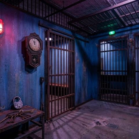 Dim jail cell set with a red caged lamp, a wall clock and keys left on a steel bench